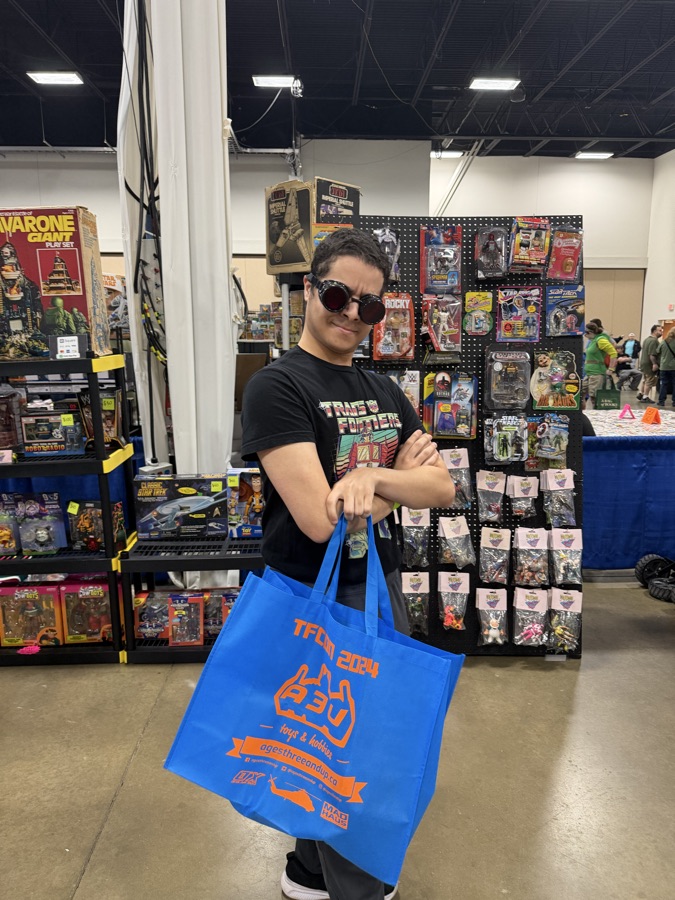 A young collector in a Transformers shirt holding a full shopping bag in front of a booth of vintage toys