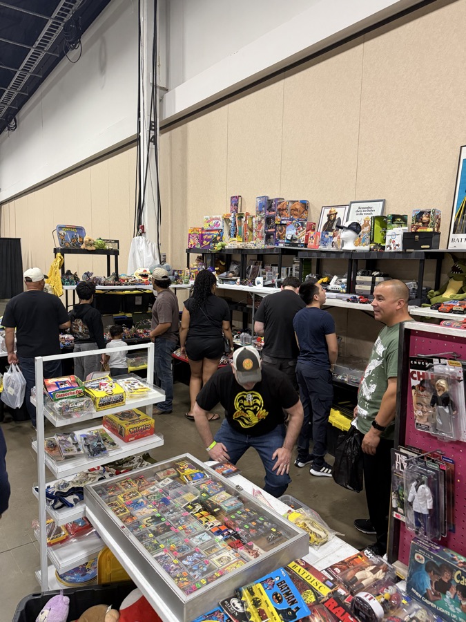 Collectors leaning over tables of vintage toys, trading cards, and action figures in a busy convention aisle